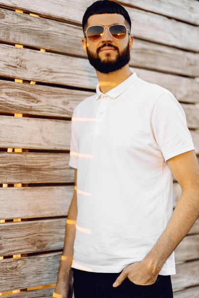 Happy young handsome man in sunglasses and with a beard, in casual outfit standing near a wooden wall, Handsome caucasian man smiling happy leaning on the wall outdoors - Photo, image