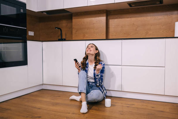 A young woman sitting on the floor in the kitchen with a phone in her hands is celebrating victory. - Photo, Image