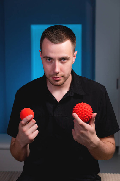 Attractive Caucasian young male masseur holding two massage balls in his hands and looking into the camera. Choice of instruments for self-massage - Foto, imagen
