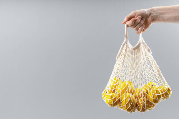 Man's hand holds string bag with lemons on solid gray background. Eco-friendly spring concept. - Fotó, kép