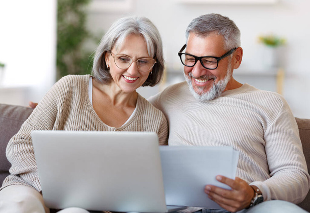Lächelndes Senioren-Paar liest Benachrichtigungsbrief mit guten Nachrichten von der Bank, während es mit Laptop sitzt - Foto, Bild
