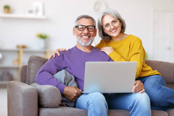 Beautiful mature family couple looking at laptop with smile on face while spending time together - 写真・画像