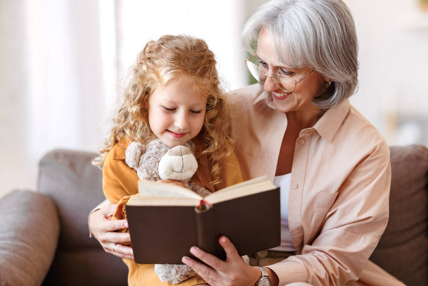 Cute little girl granddaughter reading book with positive senior grandmother - Photo, Image