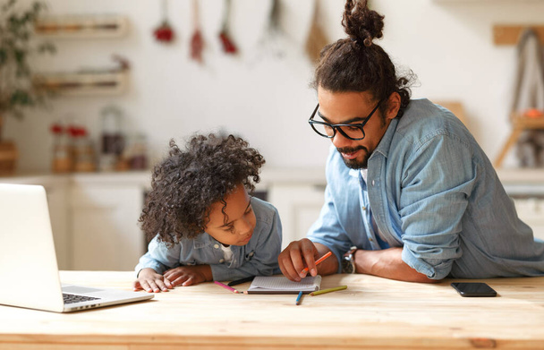 Młody afro amerykański mężczyzna ojciec wyjaśniając pracę domową do dziecka syn podczas nauki online w domu - Zdjęcie, obraz