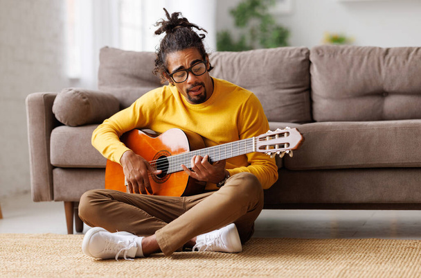 Młody, radosny, afrykański Amerykanin gra na gitarze akustycznej w domu, siedzi na podłodze w salonie - Zdjęcie, obraz
