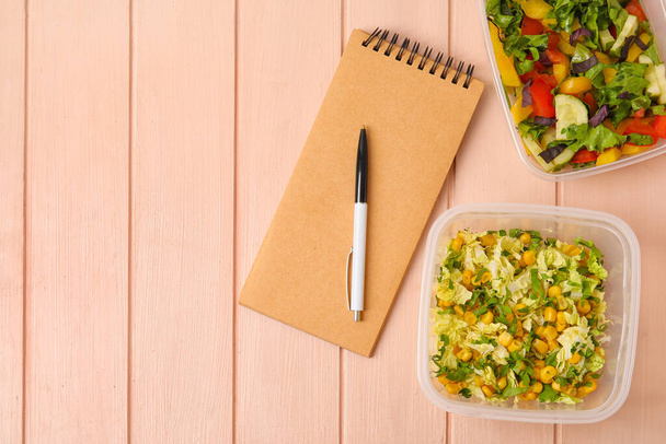 Notizbuch, Stift und Behälter mit verschiedenen Salaten auf farbigem Holzhintergrund - Foto, Bild