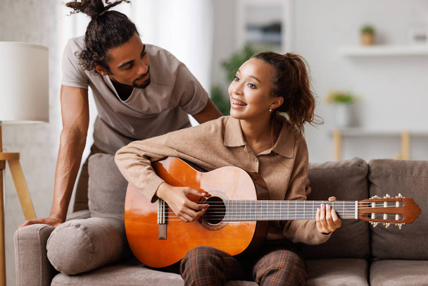 Smiling mixed race woman on sofa playing guitar for boyfriend while relaxing on weekend at home - Foto, Imagem