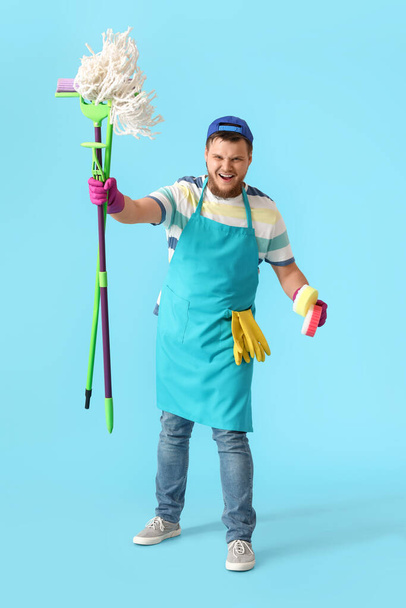 Angry young man with floor mops and sponges on blue background - Фото, изображение