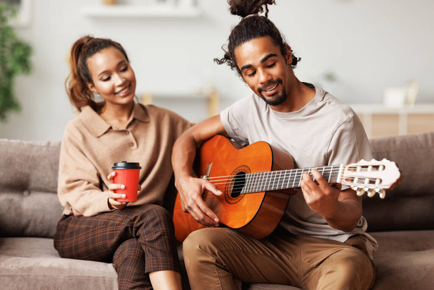 Młody uśmiechnięty Afrykanin amerykański mężczyzna muzyk gra instumentalną muzykę na gitarze dla szczęśliwej dziewczyny - Zdjęcie, obraz