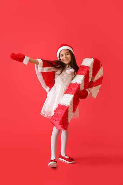 Little girl in Santa hat and with candy cane pinata on color background - 写真・画像