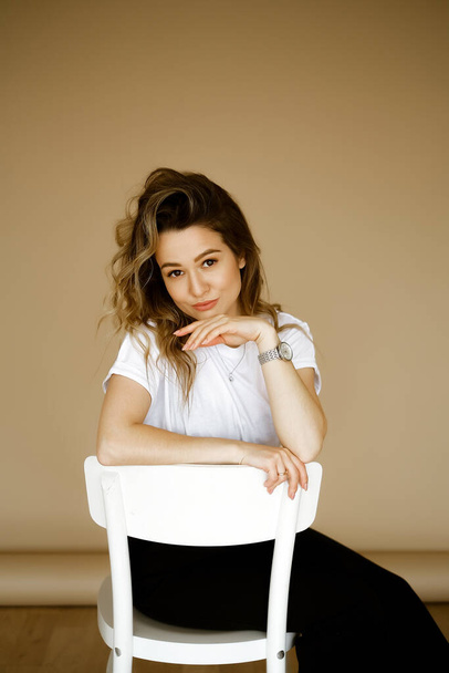 young caucasian woman in black pants and a white T-shirt sitting on a chair, portrait of a brown-eyed brunette with makeup and curly hair, lady in monochrome clothes on a light background - 写真・画像