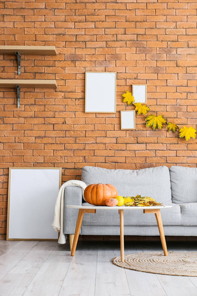 Comfortabele bank en tafel met pompoenen in de buurt van bakstenen muur met herfstbladeren en blanco frames - Foto, afbeelding