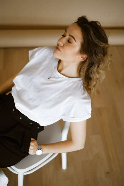 young caucasian woman in black pants and a white T-shirt sitting on a chair, portrait of a brown-eyed brunette with makeup and curly hair, lady in monochrome clothes on a light background - Foto, Imagem