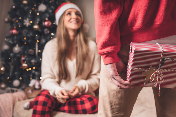 Γκρο πλαν πίσω όψη του ανθρώπου σε κόκκινο πουλόβερ κρατώντας ένα κουτί δώρου Χριστουγέννων πίσω από την πλάτη του, ενώ στέκεται κοντά διακοσμημένο χριστουγεννιάτικο δέντρο. Οικογενειακό ζευγάρι στις διακοπές των Χριστουγέννων. - Φωτογραφία, εικόνα