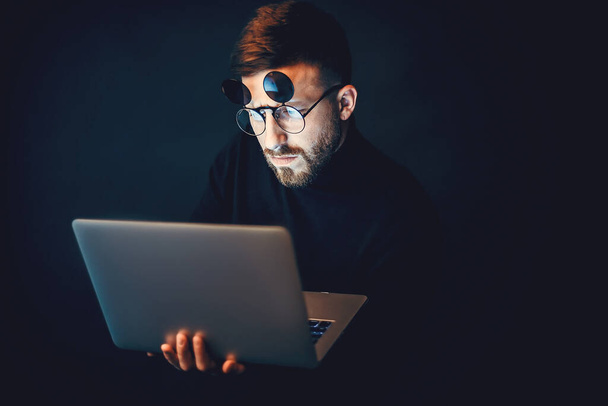 Fiatal szakállas hipster férfi visel szemüveg használata közben laptop felett fekete stúdió háttér  - Fotó, kép