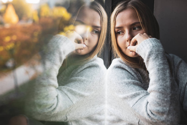 Depressed and anxious young woman sitting by a large window, feeling blue, sad, uncertain - Fotografie, Obrázek