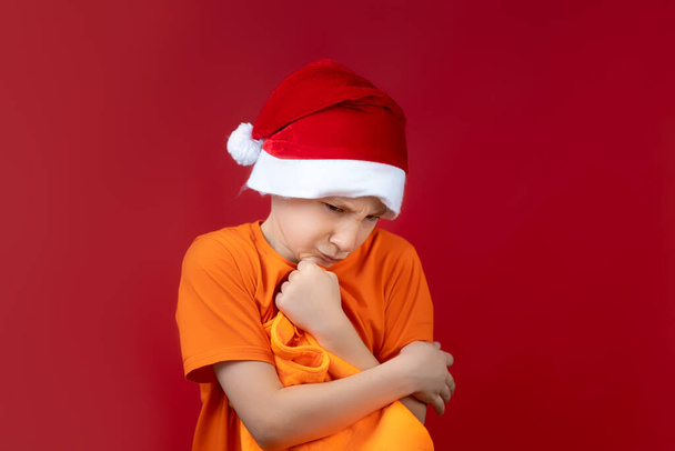 um menino em uma camisa amarela fica em um fundo vermelho usando um chapéu de Papai Noel segurando um saco de presente em suas mãos - Foto, Imagem