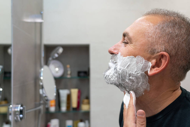 Érett, jóképű férfi borotvahabot hord az arcára, hogy levágja a szakállát és a bajuszát borotvával. - Fotó, kép