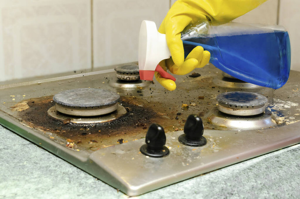 Cleaning dirty gas stove from grease, food leftovers deposits. womans hand in protective glove with sponge rag and detergent in bottle washing kitchen stove. home cleaning service concept - Fotografie, Obrázek