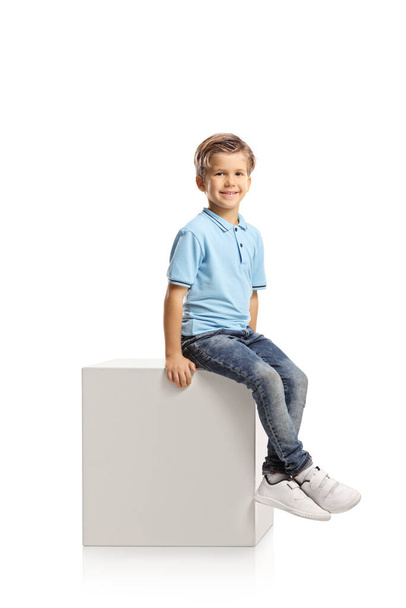 Smiling caucasian blond boy sitting on a white cube isolated on white background - Foto, Imagem