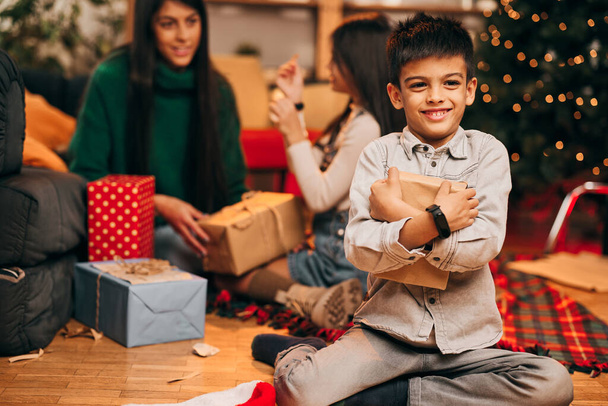 Милый маленький мальчик, сидящий дома и обнимающий рождественский подарок, который он хотел. Новогодние и рождественские праздники. - Фото, изображение