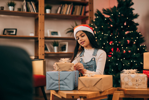 Cute little girl with Santa hat on her head kneeling next to a pile of Christmas presents on Christmas eve. - Photo, Image