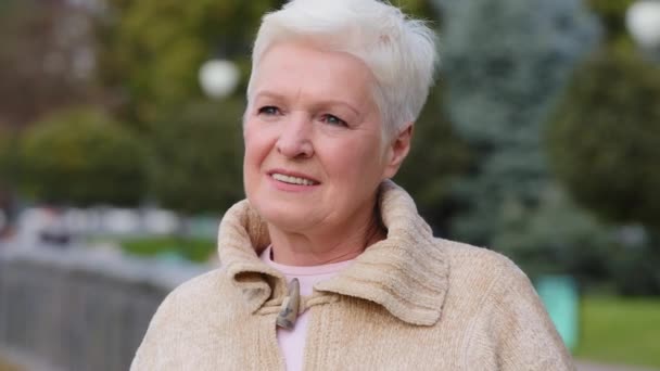 Closeup portrait smiling mature lady in casual clothes standing posing outdoors looking aside turns head at camera. Calm attractive benevolent woman of retirement age with gray stylish short hair - Footage, Video