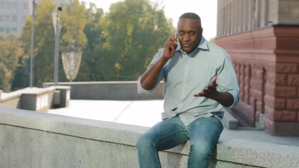 Беспокоится, что афроамериканец в рубашке и джинсах держит мобильный телефон. Разочарованный клиент пенсионного возраста разговаривает с обслуживанием, сидя на открытом воздухе - Кадры, видео