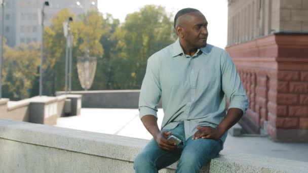 Estresado hombre afroamericano frustrado de edad de jubilación en ropa casual camisa y jeans sentado al aire libre esperando a cliente o amigo tarde para la reunión, mirando ansiosamente a la pantalla del teléfono inteligente - Metraje, vídeo