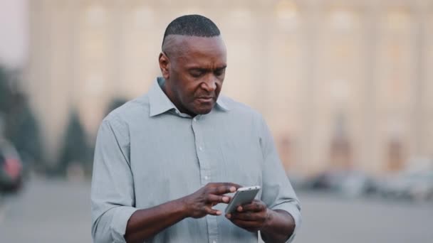 Adult mature black man holding smartphone looking at phone screen look for address with electronic map in city. Elderly businessman African American male professional finding office building outdoors - Footage, Video