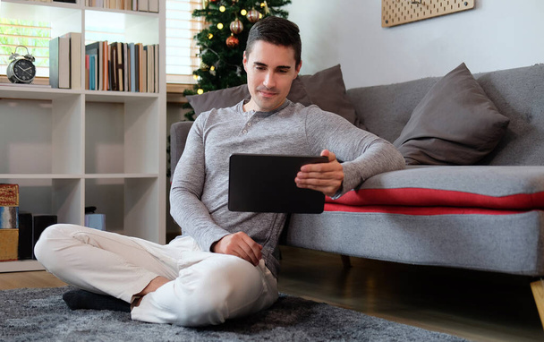 Jonge man in casual kleding zittend in de woonkamer en met behulp van digitale tablet. - Foto, afbeelding
