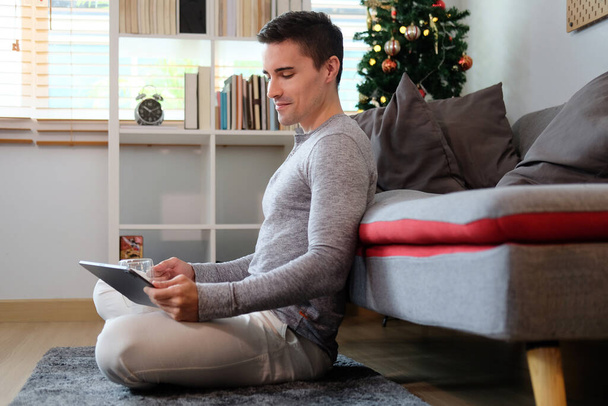 Handsome man in casual clothes sitting in living room and surfing internet with digital tablet. - Photo, Image