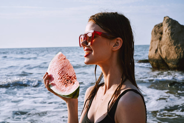 cheerful woman in black swimsuit eating watermelon on the beach - Фото, зображення