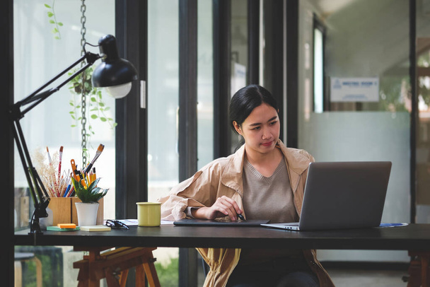 Pewna siebie twórcza azjatycka kobieta pracuje z laptopem w biurze. - Zdjęcie, obraz