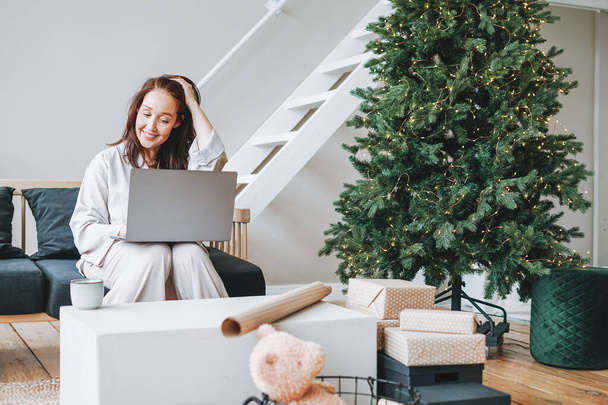 Junge Erwachsene vierzig Jahre Frau mit dunklen Haaren im gemütlichen Pyjama mit Geschenk-Box sitzen und mit Laptop im Wohnzimmer mit Weihnachtsbaum zu Hause - Foto, Bild
