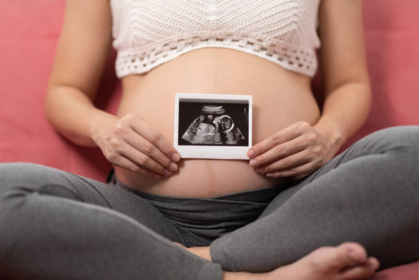 Close up Happy Pregnant Woman holding ultrasound image stroking big belly with love isolated on couch,Pregnancy of young woman enjoy with future life,Motherhood and Pregnant Concept - Фото, зображення
