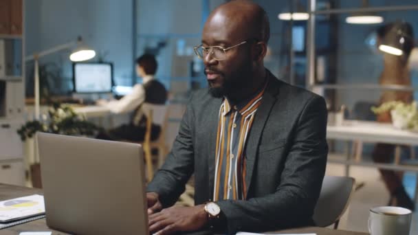 African American businessman in glasses and formal outfit working laptop at office desk in evening - Materiał filmowy, wideo