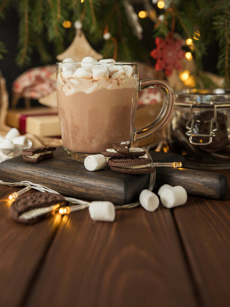 Ζεστή σοκολάτα με ζαχαρωτό σε γυάλινο κύπελλο πάνω σε ξύλινο τραπέζι κάτω από το χριστουγεννιάτικο δέντρο. Ζεστό ρόφημα και χριστουγεννιάτικο δέντρο. Χριστουγεννιάτικη κάρτα χαιρετισμού. - Φωτογραφία, εικόνα