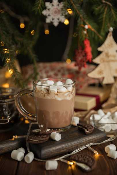 Hot chocolate with marshmallow  in a glass cup on a wooden table under the Christmas tree . Hot beverage and Christmas tree . Christmas greeting card. - Фото, изображение