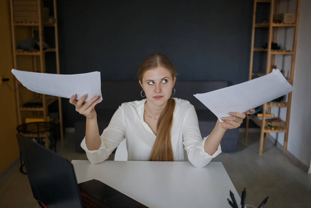 A young successful employee of a business woman in a white shirt in surprise holds white sheets of paper, documents in her hands, sits at a white desk workplace with a laptop computer in a modern office indoors. The concept of career achievement, suc - Fotoğraf, Görsel