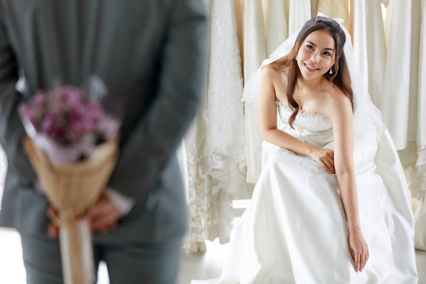 Asiatische junge schöne langhaarige Braut in weißem Brautkleid mit durchsichtigem Spitzenschleier sitzt lächelnd und wartet auf den Bräutigam im grauen Anzug, der den Blumenstrauß hinter dem Rücken versteckt, um in der Garderobe zu überraschen. - Foto, Bild