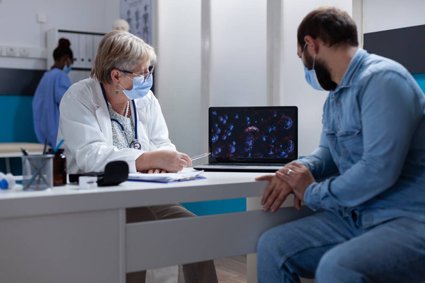 Ασθενής και ειδικός με μάσκα προσώπου κοιτάζοντας απεικόνιση του ιού στο laptop - Φωτογραφία, εικόνα