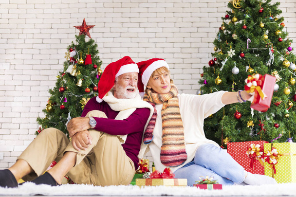 Starsza kaukaska para świętuje Boże Narodzenie razem w szczęściu i ekscytacji w domu z czerwonym kapeluszem Mikołaja i choinką - Zdjęcie, obraz