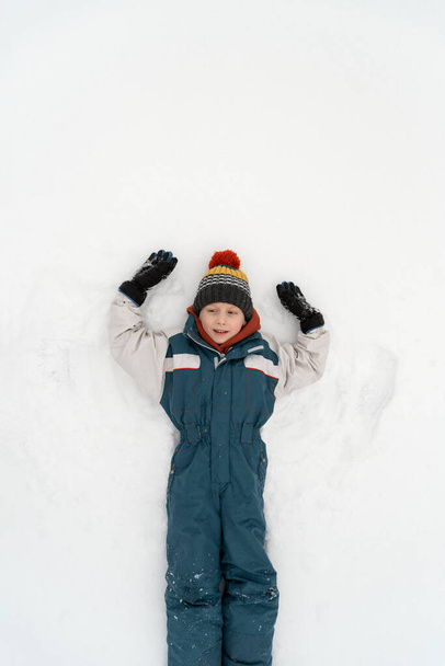 Grappige jongen in ski jumpsuit ligt in heldere witte sneeuw. Kind maakt sneeuwengel. Bovenaanzicht. Verticaal frame. - Foto, afbeelding