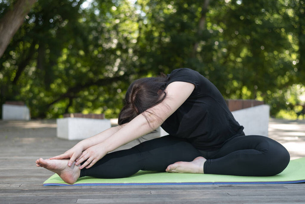 Young beautiful flexible girl makes a lean forward with her hands from a sitting position. Side view. On a green mat. - Photo, Image