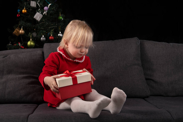La bambina apre la confezione regalo seduta sul divano sullo sfondo di un albero di Natale. La vigilia di Natale. Regali di Capodanno - Foto, immagini