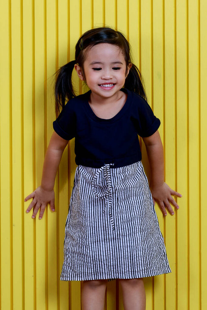 Portrét studio záběr malé roztomilé krásné copánky účes předškolačka šťastná dívka model nosí černou košili a bílý proužek sukně stojí s úsměvem pózuje před žlutou stěnou pozadí. - Fotografie, Obrázek