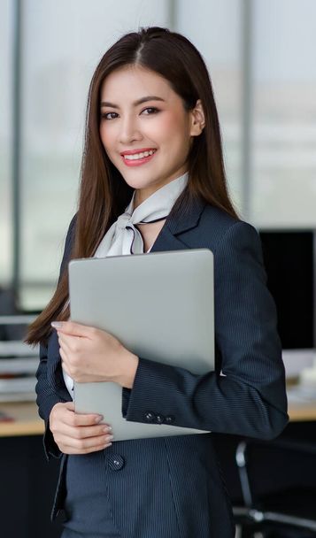 Porträt einer jungen attraktiven asiatischen Büroangestellten in formalen Business-Anzügen, die im Büro in die Kamera lächelt, mit verschwommenem Büro als Hintergrund. - Foto, Bild