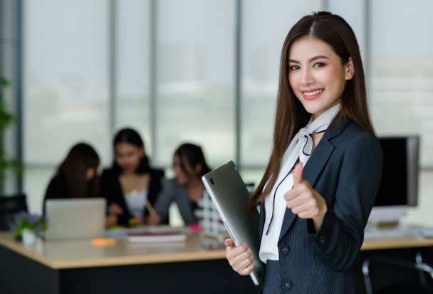 Portrait of young attractive Asian female office worker in formal business suits  smiling at camera in office with blurry colleagues sitting in office as background. - Fotografie, Obrázek