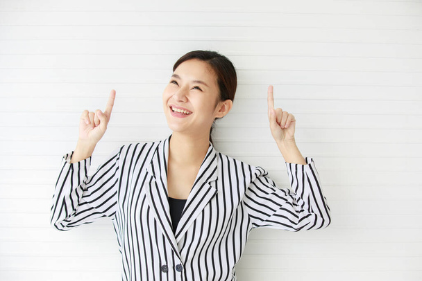 Portrait shot of Asian young confident happy female businesswoman wears black white stripe casual suit stand in front wall smiling hold hands point fingers and look up above on empty copy space. - Photo, image
